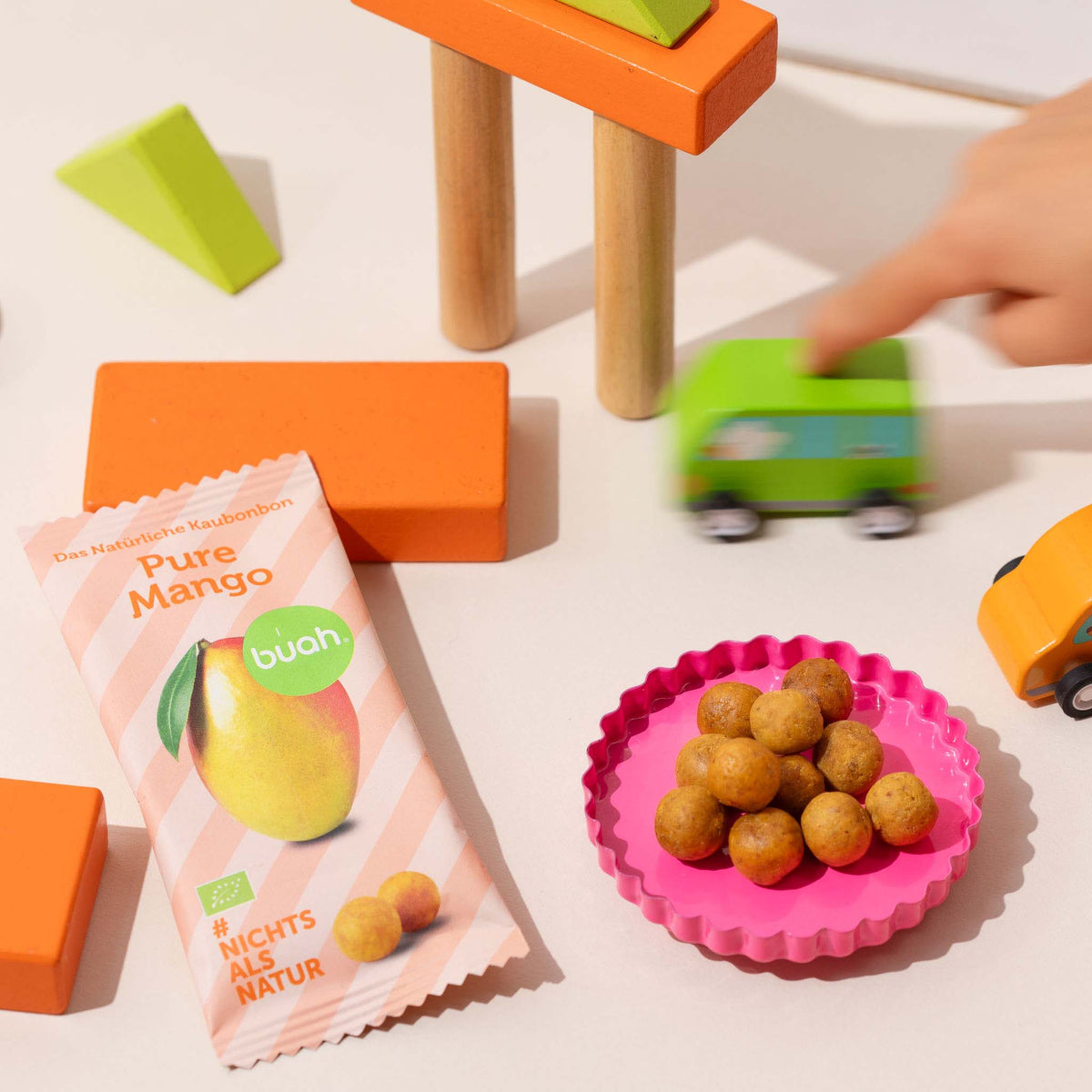 buah Kaubonbon Mango Packung neben Kaubonbons in Schale und Holzspielzeug im Hintergrund