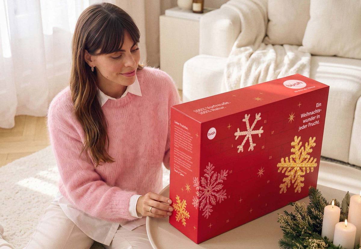 Eine lächelnde Frau im rosa Pullover sitzt auf dem Boden an einem Couchtisch und betrachtet voller Vorfreude den roten buah Adventskalender. Sie ist dabei, eines der Türchen zu öffnen.