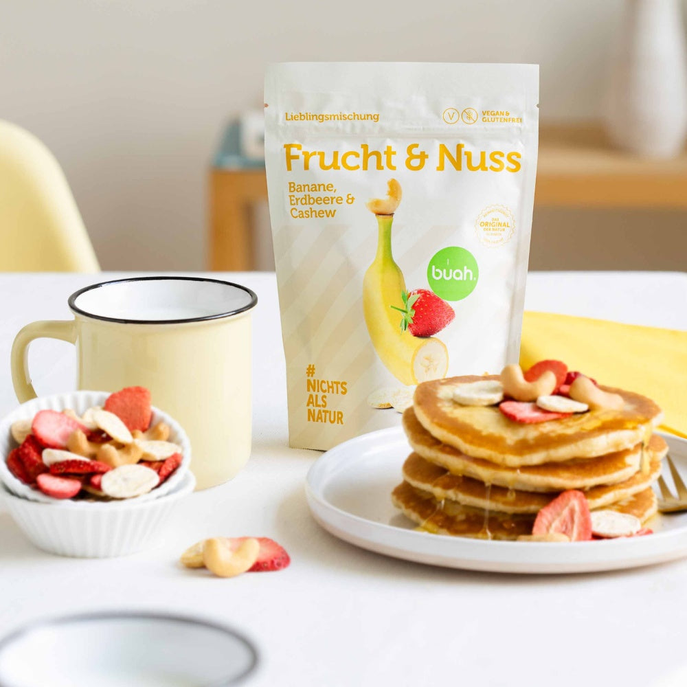 Packung buah Frucht & Nuss Mischung steht neben Pancakes, dekoriert mit buah Bananen, Erdbeeren und Cashews