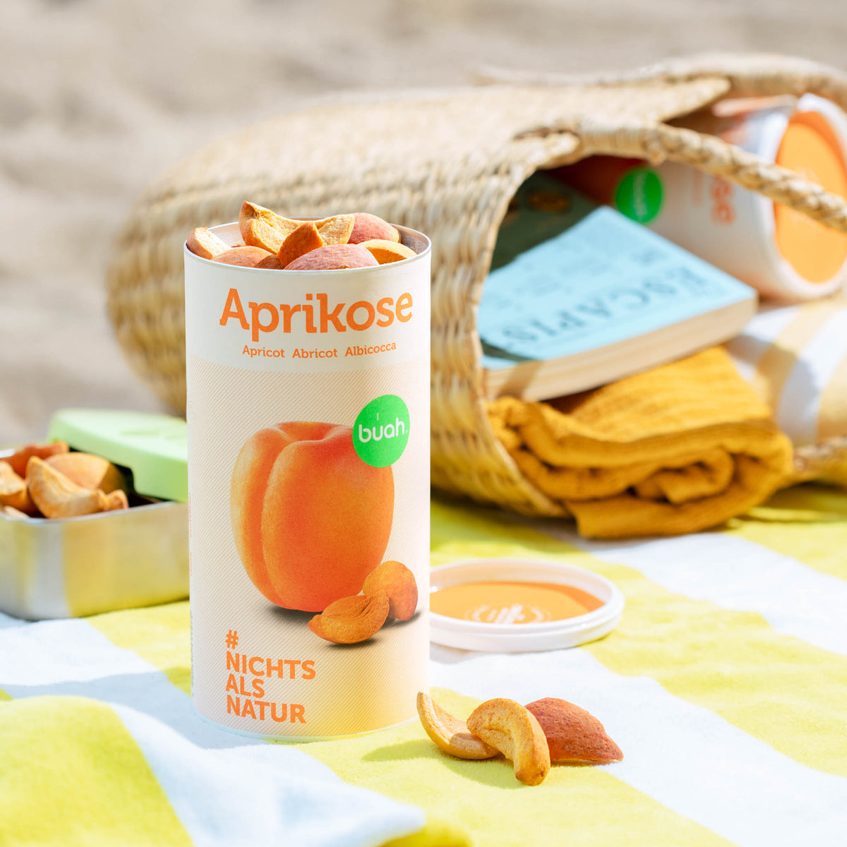 Dose buah Aprikose steht auf einem gelb gestreiftem Handtuch am Strand, vor der Dose liegen gefriergetrocknete Aprikosen Schnitze