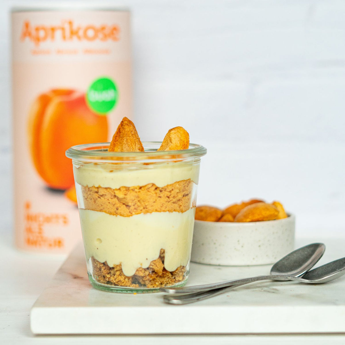 Dessertglas mit Schichten aus Joghurt und buah gefriergetrockneter Aprikose steht vor einer Dose buah Aprikose, daneben zwei Loeffel