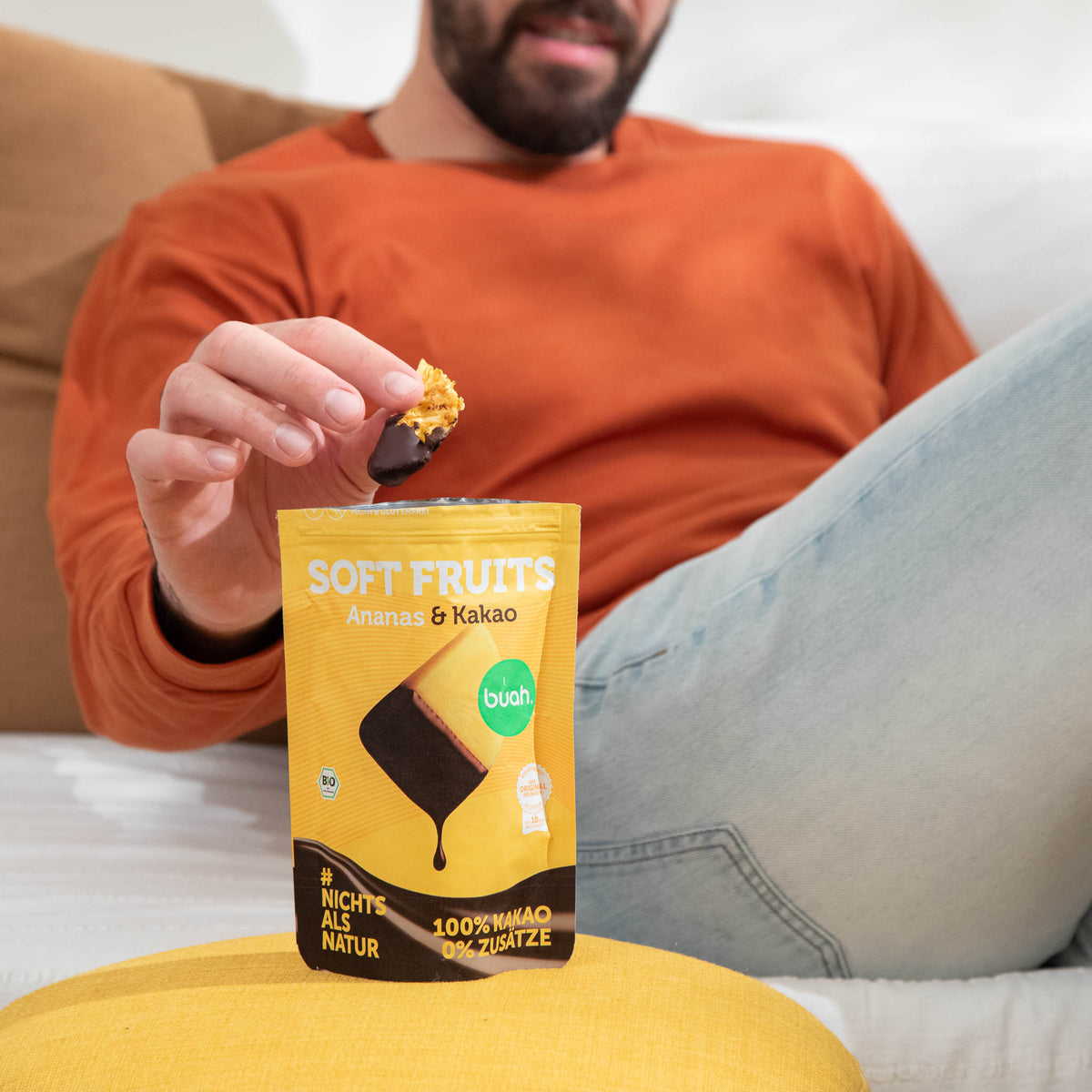 Ein lächelnder Mann im orangefarbenen Pullover sitzt entspannt auf einem weißen Sofa. Er isst ein schokoliertes buah Soft Fruit Ananas-Stück und hat den gelben Beutel der Sorte Ananas & Kakao auf seinem Schoß liegen.