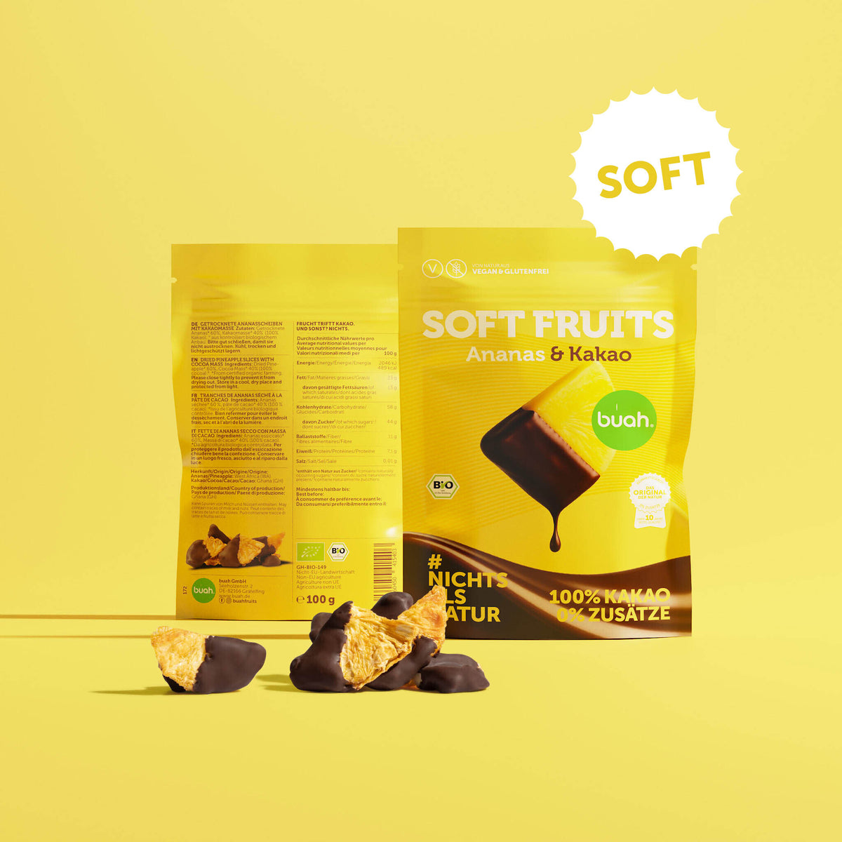 Ein gelber Beutel buah Soft Fruits der Sorte Ananas & Kakao steht vor einem gelben Hintergrund. Dahinter ist die RĂĽckseite der Verpackung zu erkennen. Davor liegen einige softe, mit Schokolade ĂĽberzogene Ananas-StĂĽcke.