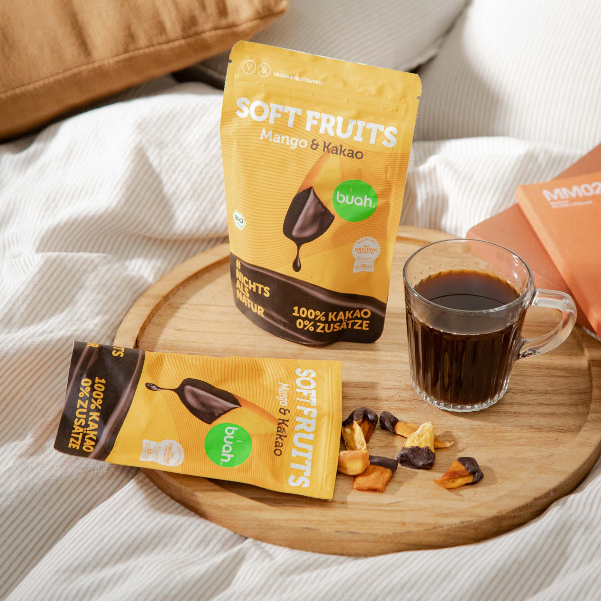 Ein gemütliches Arrangement auf einem Bett. Ein geöffneter Beutel buah Soft Fruits Mango & Kakao liegt auf einem runden Holztablett, daneben eine Glastasse mit Kaffee und verstreute schokolierte Mango-Stücke.