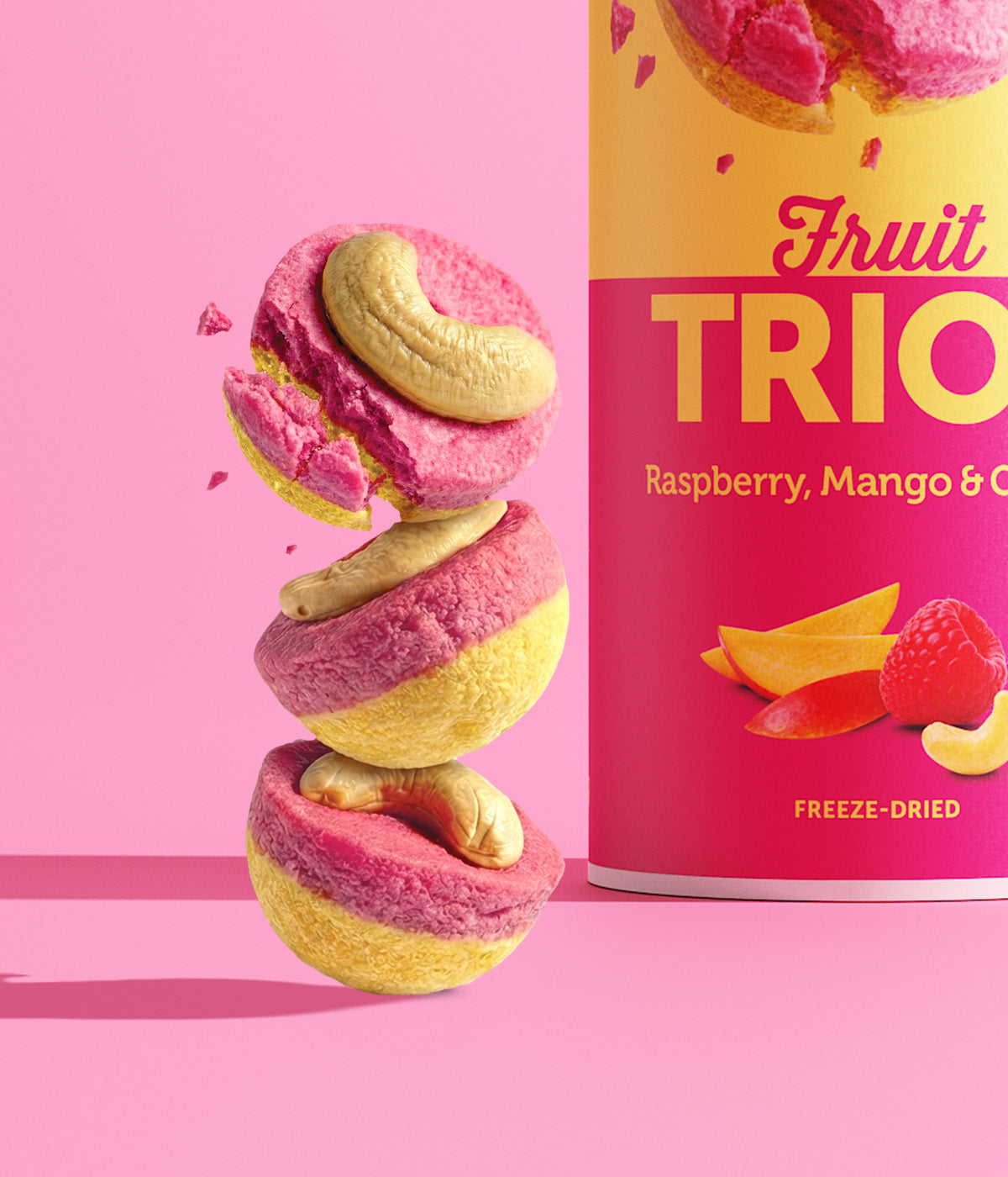 Ein Turm aus drei runden buah Fruit TRIOS StĂĽcken der Sorte Raspberry, Mango & Cashew vor pinkem Hintergrund. Das oberste, pink-gelbe FruchtstĂĽck zerbricht in der Luft. Auf jedem StĂĽck liegt ein Cashewkern.