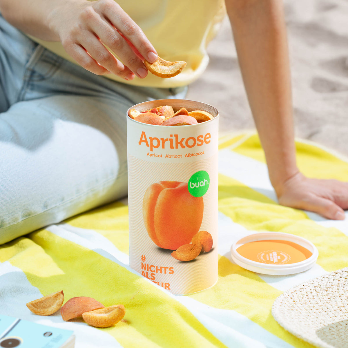 Person nimmt buah Aprikose aus der Dose und sitzt auf einem gelb gestreiftem Handtuch am Strand 