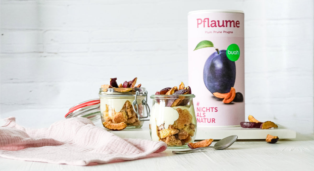 Dessert aus Crumble, Joghurt und buah gefriergetrockneter Pflaume in einem Glas, steht auf einem Tisch mit einer Dose buah Pflaumen daneben