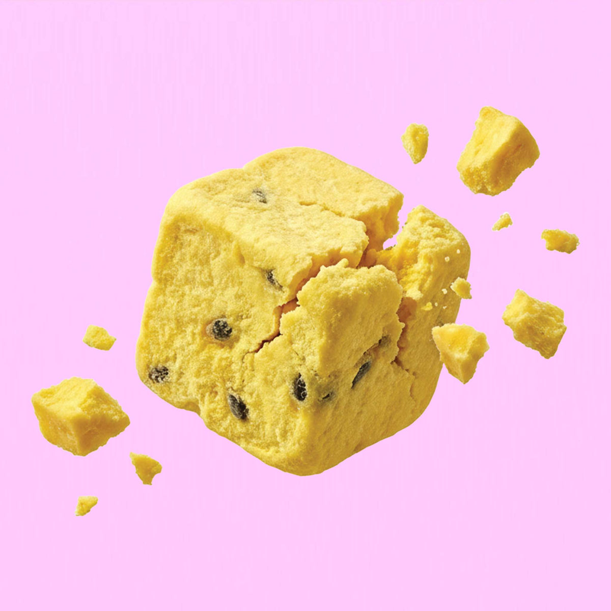 Ein quadratischer, buah gefriergetrockneter Fruchtwürfel der Sorte Mango & Maracuja zerbricht in der Luft vor einem rosafarbenen Hintergrund. Kleine gelbe Stückchen und dunkle Maracujakerne fliegen davon.