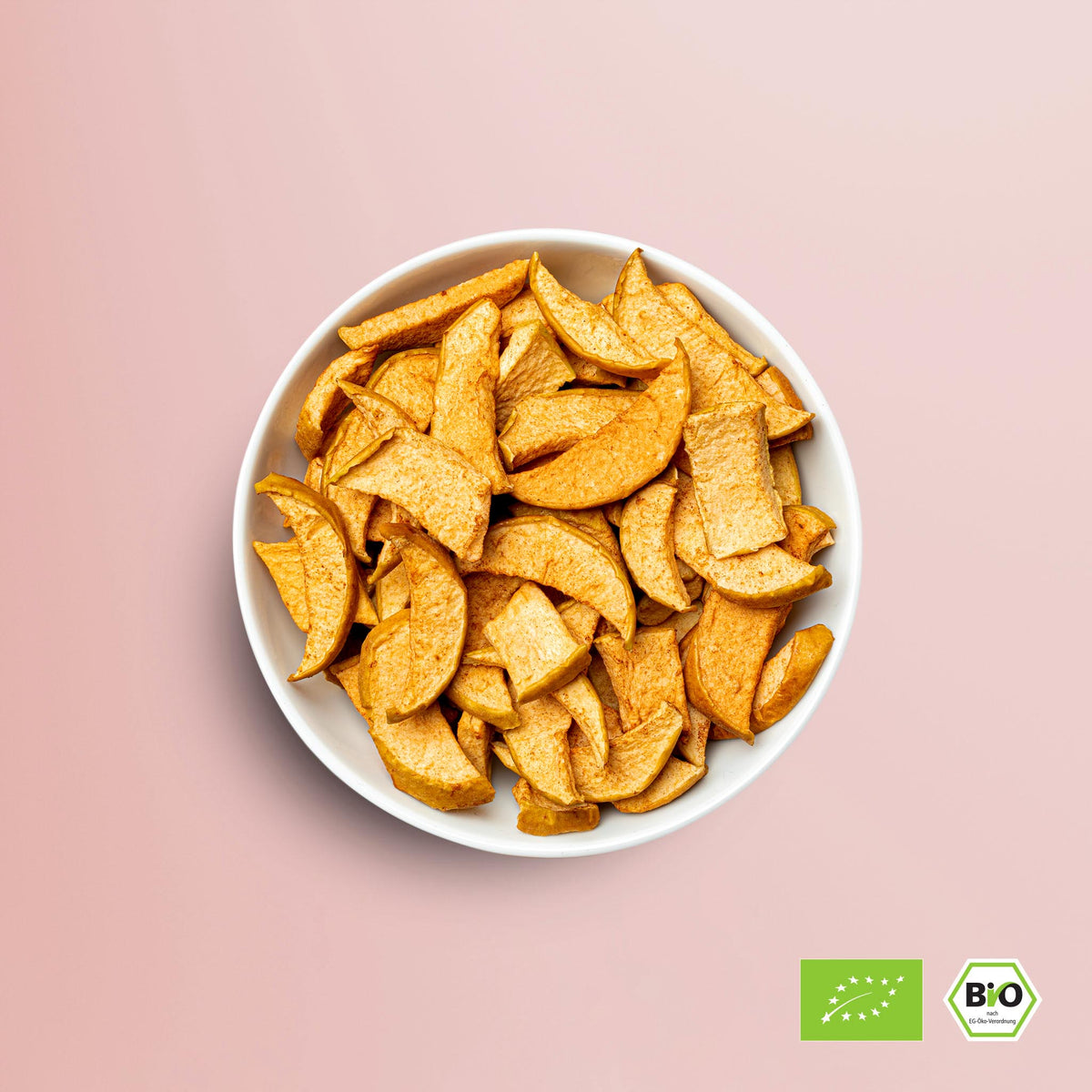 Weiße Bowl mit buah gefriergetrocknetem ApfelZimt in gelblicher Farbe mit braunem Staub, vor rosa Hintergrund, BIO Logo in der Ecke der Abbildung
