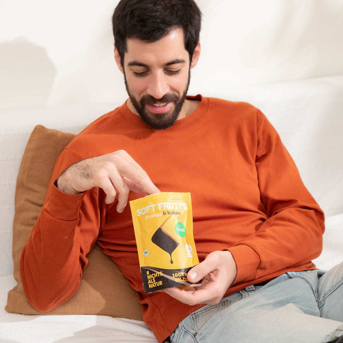 Ein Mann im orangefarbenen Pullover sitzt auf einem weißen Sofa und greift in einen geöffneten gelben Beutel buah Soft Fruits der Sorte Ananas & Kakao.
