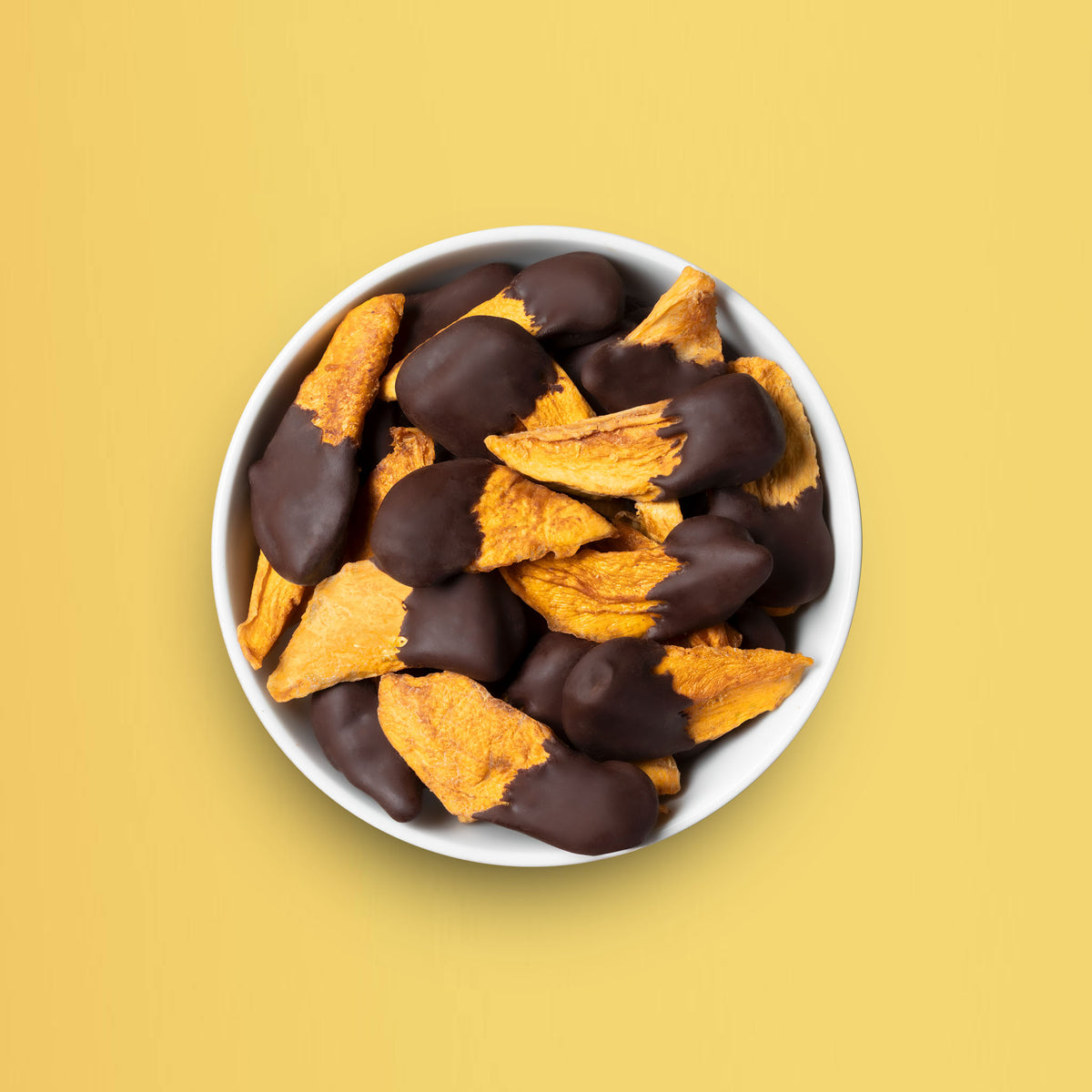 Eine weiße Schale gefüllt mit soften buah Mango-Stücken in dunkler Schokolade vor einem hellgelben Hintergrund.