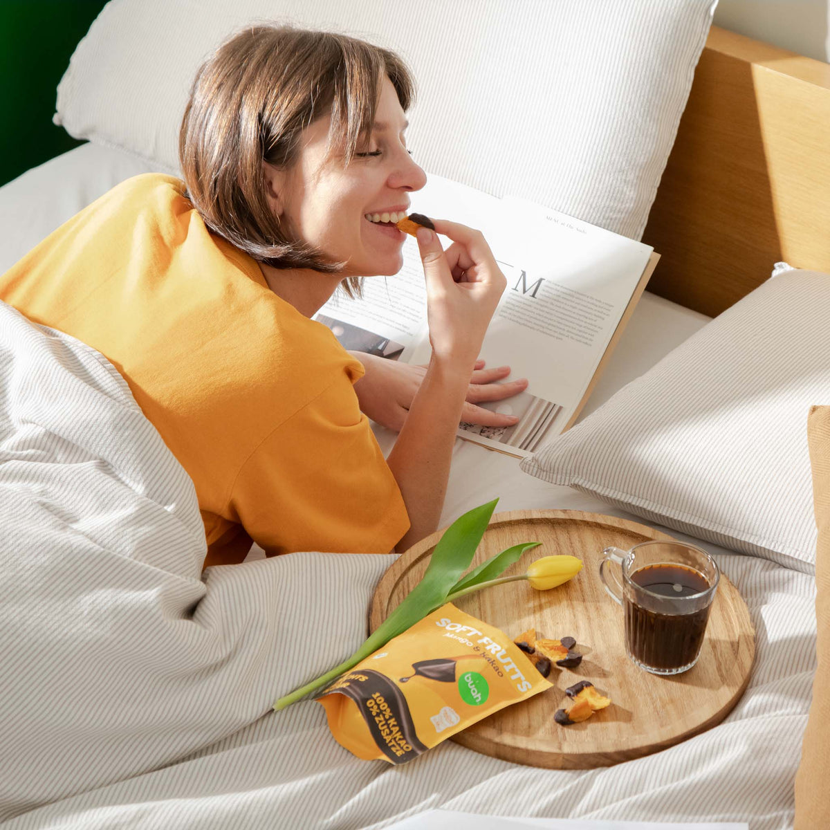 Eine lächelnde Frau liegt im Bett, liest ein aufgeschlagenes Magazin und isst genussvoll ein schokoliertes buah Mango-Stück. Auf dem Bett vor ihr steht ein Holztablett mit dem Snack-Beutel, Kaffee und einer gelben Tulpe.