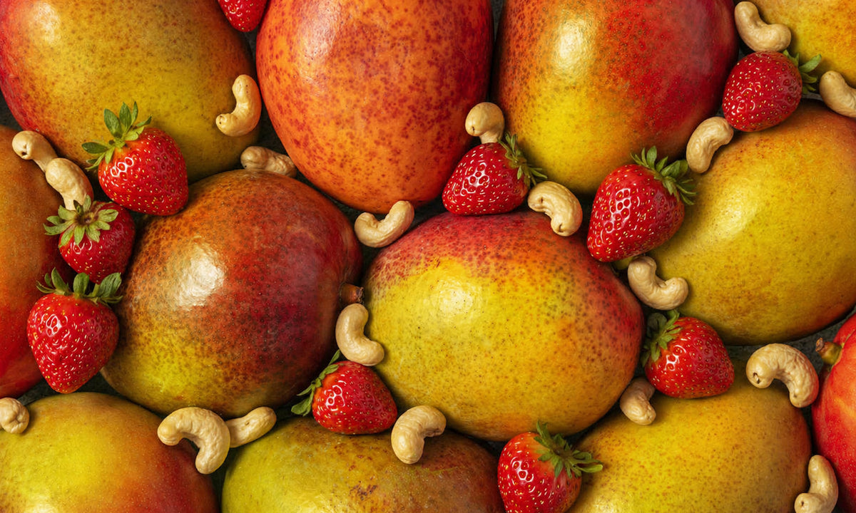 Eine farbenfrohe Nahaufnahme von vielen frischen, ganzen Mangos. Dazwischen liegen frische Erdbeeren und helle Cashewkerne.